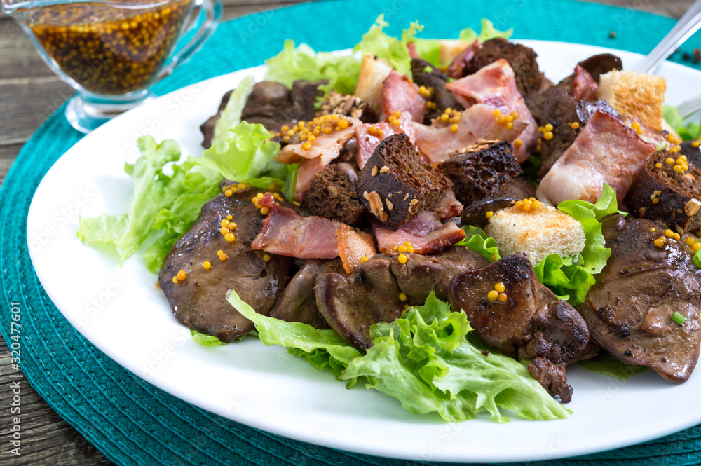 Warm healthy salad of chicken liver, rye croutons, smoked bacon, green salad and mustard sauce in a white plate on a wooden background. Close up Warm healthy salad of chicken liver, rye croutons, smoked bacon, green salad and mustard sauce in a white plate on a wooden background. Close up