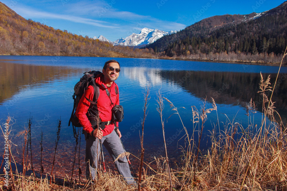 A middle-aged man in a red jacket and glasses with a backpack for tourism on the background of the lake which reflects the snow-capped mountains. A middle-aged man in a red jacket and glasses with a backpack for tourism on the background of the lake which reflects the snow-capped mountains.