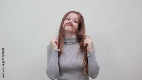 young beautiful red haired woman in gray sweater made her hair an imitation of man's moustache with silly and funny expression on face