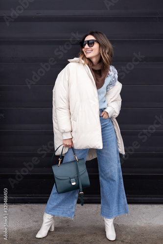 European model girl in a beige oversized down jacket, knitted sweater, flared jeans with a handbag and glasses posing on the street. Life style