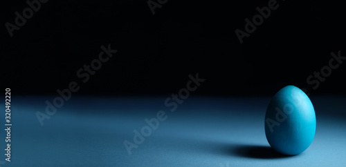 Moody easter banner concept in classic blue color. Blue cyan colored egg on black background. Selective focus. Light and shadow. Copy space. Minimal.