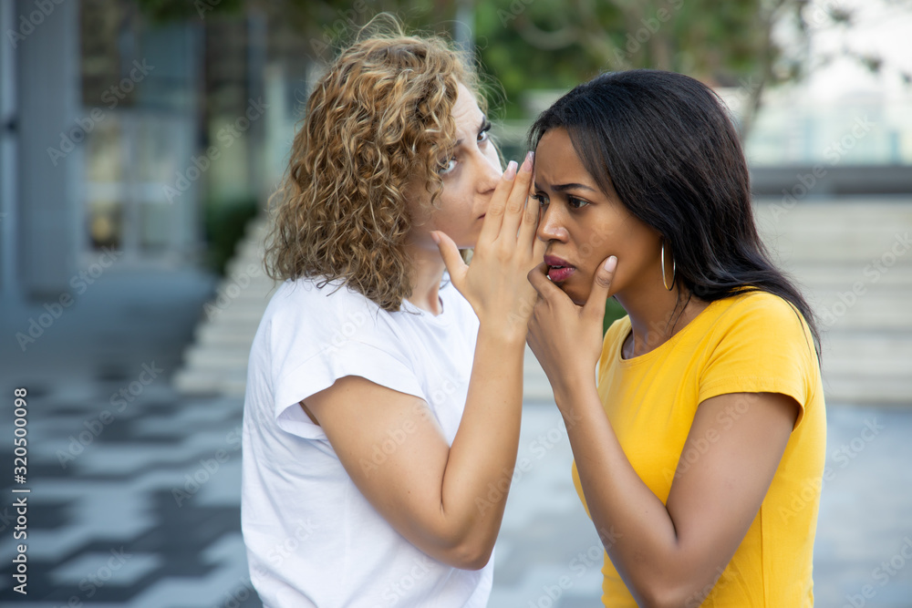 black and white women gossiping and whispering; concept of rumor, gossip, hearsay, bad news, social media network fake news, news update; caucasian and african woman real people model black and white women gossiping and whispering; concept of rumor, gossip, hearsay, bad news, social media network fake news, news update; caucasian and african woman real people model