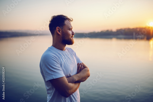 Картината върху платно Handsome bearded blond caucasian man standing on cliff with arms crossed and looking at beautiful sunset