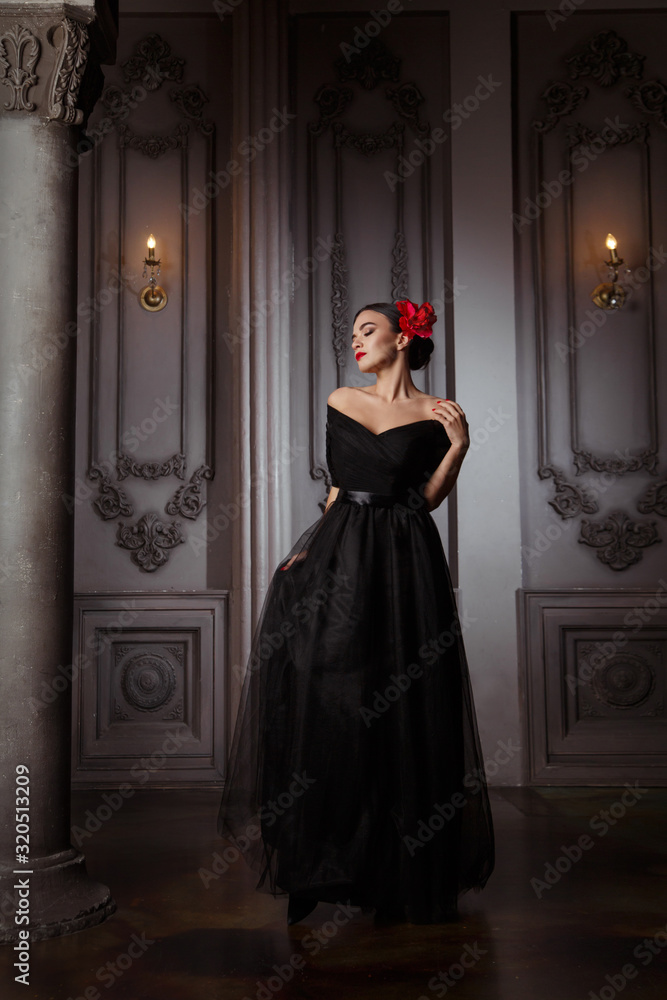 Beautiful brunette girl in a long black dress and with a red flower in a hairstyle in the palace.