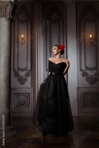 Beautiful brunette girl in a long black dress and with a red flower in a hairstyle in the palace.