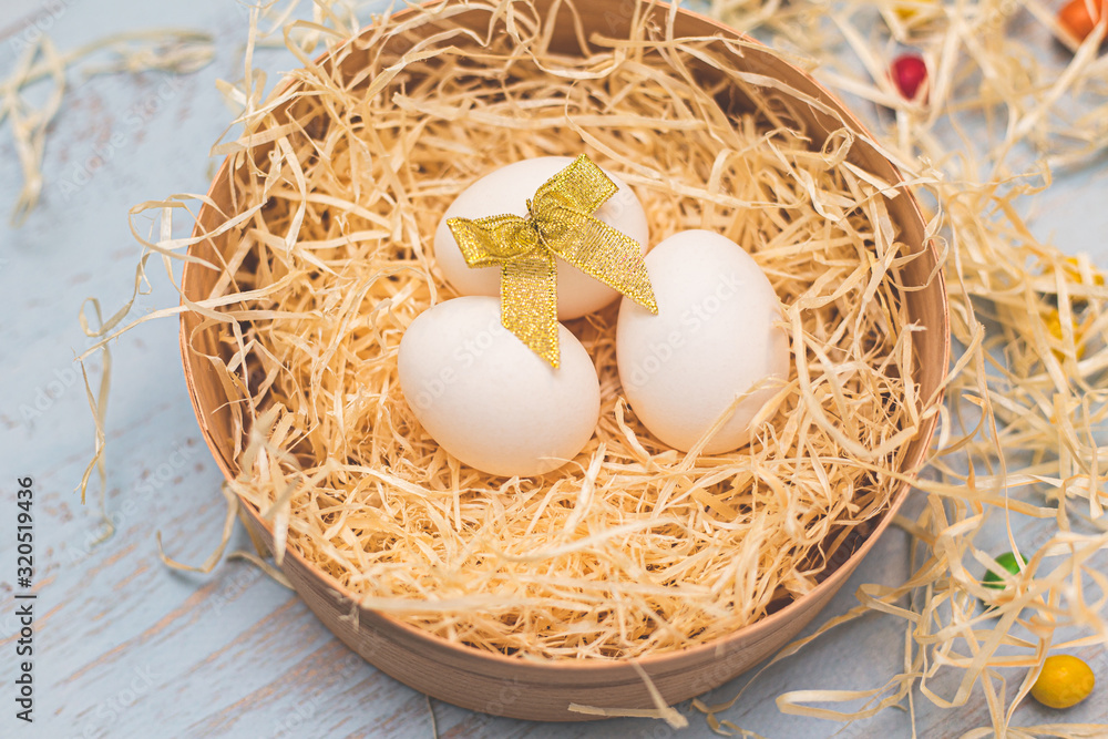 Three white eggs with a golden bow and colorful round sweets in a hay nest in a box. Top side view on a light blue wooden background. Copyspace for the wording. Easter holidays concept. Three white eggs with a golden bow and colorful round sweets in a hay nest in a box. Top side view on a light blue wooden background. Copyspace for the wording. Easter holidays concept.