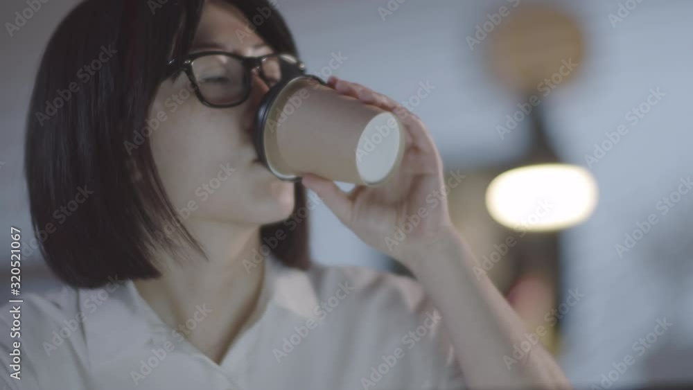 Tilt down shot of Asian businesswoman in glasses drinking coffee from to-go cup and working on laptop in dark office in evening Tilt down shot of Asian businesswoman in glasses drinking coffee from to-go cup and working on laptop in dark office in evening