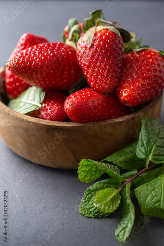 Top view of strawberries in wooden bowl and mint branch, with selective focus, on gray background in vertical