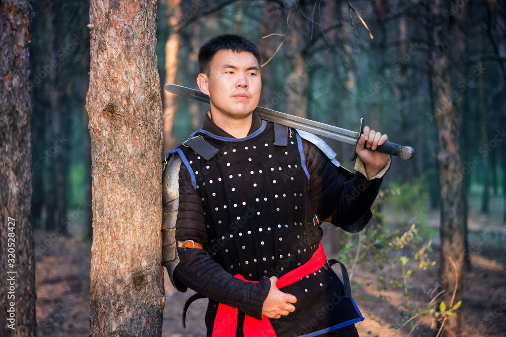 Warrior in black armor with a sword in his hands is leaning against a tree in a gloomy enchanted, damned forest.