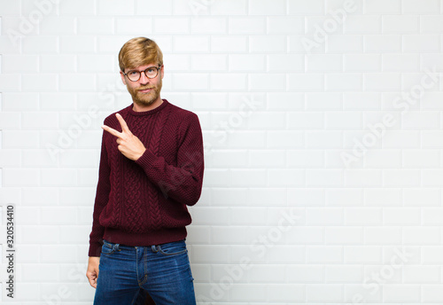 young blonde man feeling happy, positive and successful, with hand making v shape over chest, showing victory or peace agaist vintage tiles wall photo