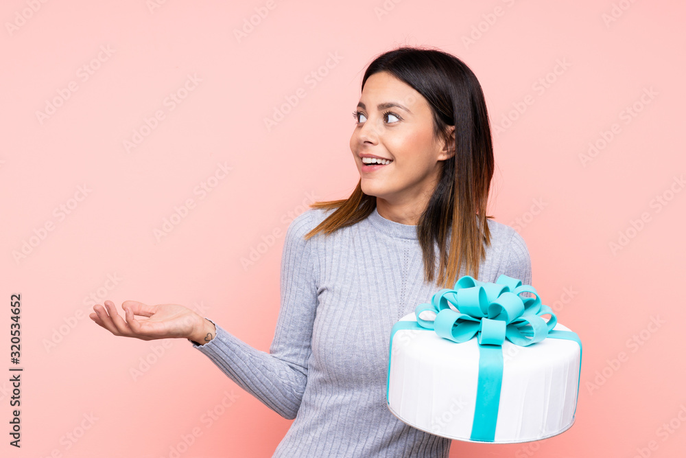 Woman holding a big cake over isolated pink background with surprise facial expression Woman holding a big cake over isolated pink background with surprise facial expression