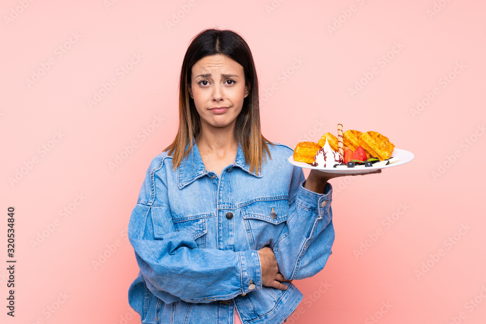 Brunette woman holding waffles over isolated pink background with sad expression