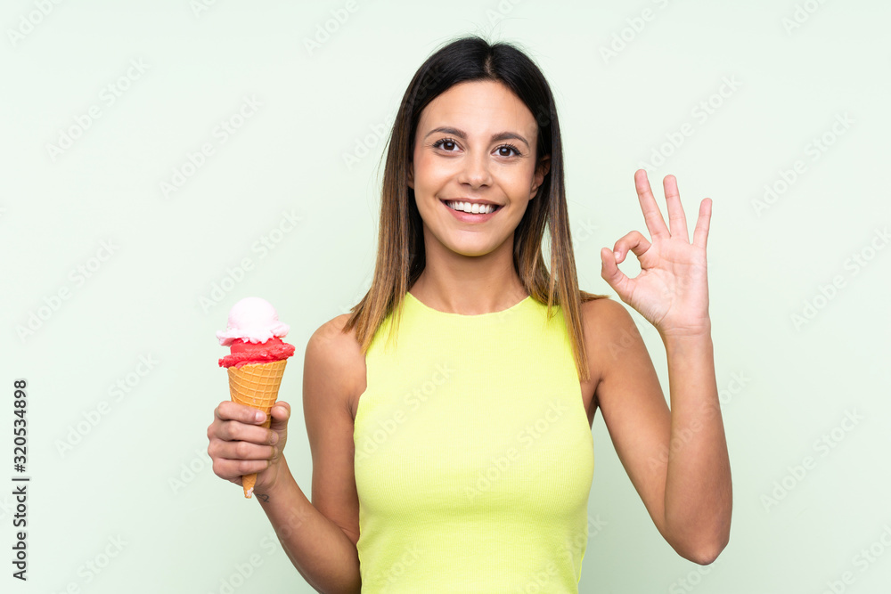 Brunette woman holding a cornet ice cream over isolated green background showing ok sign with fingers Brunette woman holding a cornet ice cream over isolated green background showing ok sign with fingers