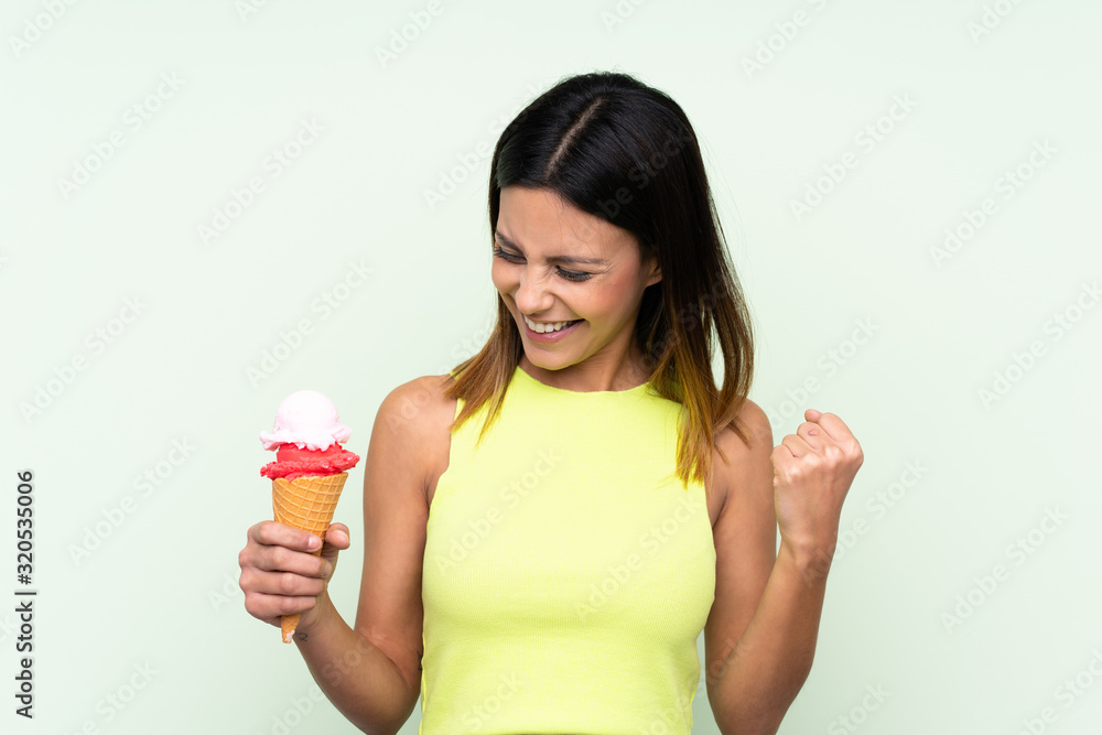 Brunette woman holding a cornet ice cream over isolated green background celebrating a victory Brunette woman holding a cornet ice cream over isolated green background celebrating a victory