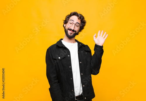 young crazy handsome man smiling happily and cheerfully, waving hand, welcoming and greeting you, or saying goodbye against orange wall photo