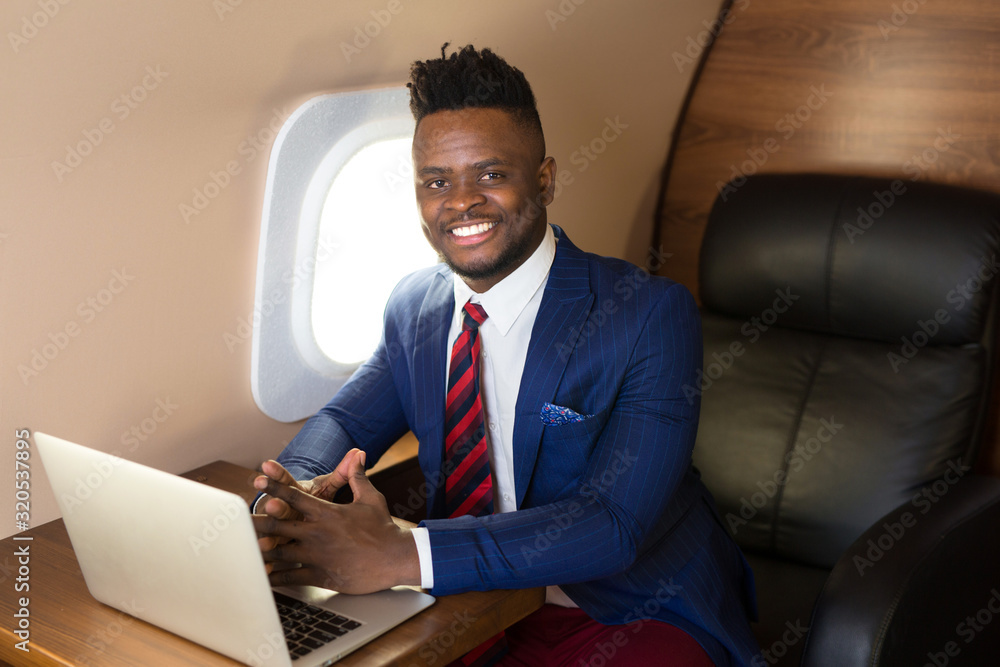 handsome african young man in suit in private jet cabin with laptop