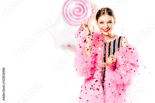 smiling girl in a striped leotard posing with eyes open while standing on a white background. young woman in a pink fur coat celebrates her birthday, has fun with golden confetti and inflatable balloo photo