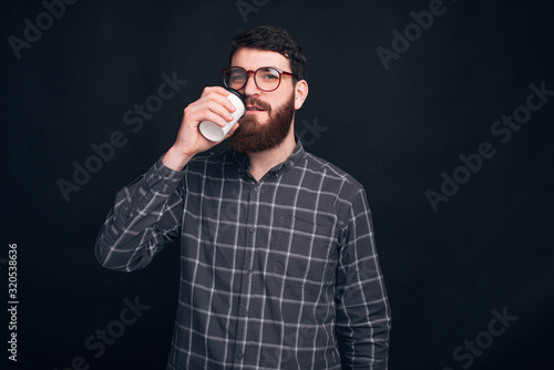 Young man wearing glasses and a nice beard is drinking something from a take away cup.