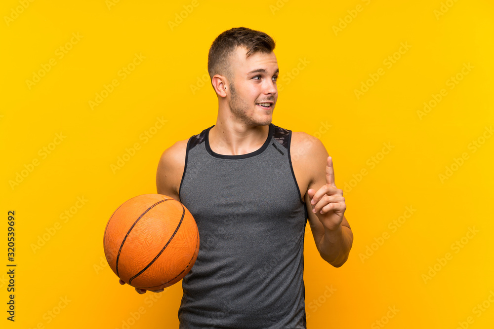 Young handsome blonde man holding a basket ball over isolated yellow background intending to realizes the solution while lifting a finger up