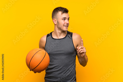Young handsome blonde man holding a basket ball over isolated yellow background intending to realizes the solution while lifting a finger up © luismolinero