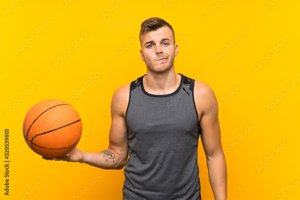 Young handsome blonde man holding a basket ball over isolated yellow background with sad expression Young handsome blonde man holding a basket ball over isolated yellow background with sad expression