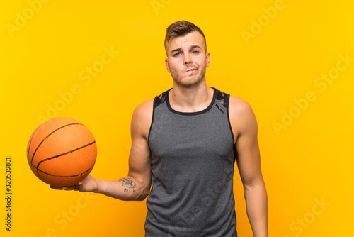 Young handsome blonde man holding a basket ball over isolated yellow background with sad expression