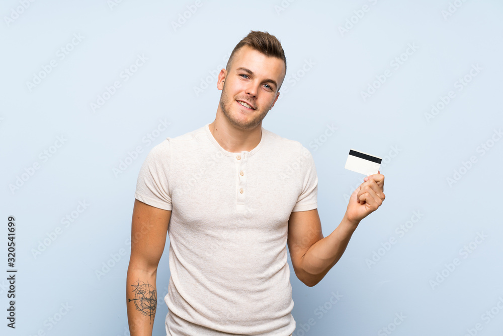 Young handsome blonde man over isolated blue background holding a credit card Young handsome blonde man over isolated blue background holding a credit card