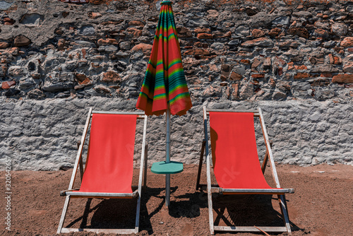 View of two inviting empty orange wooden chairs for sunbathing and a table with ditto parasol, standing in the sand on the beach in Italy, as a background a stone wall with large yellow boulders. photo