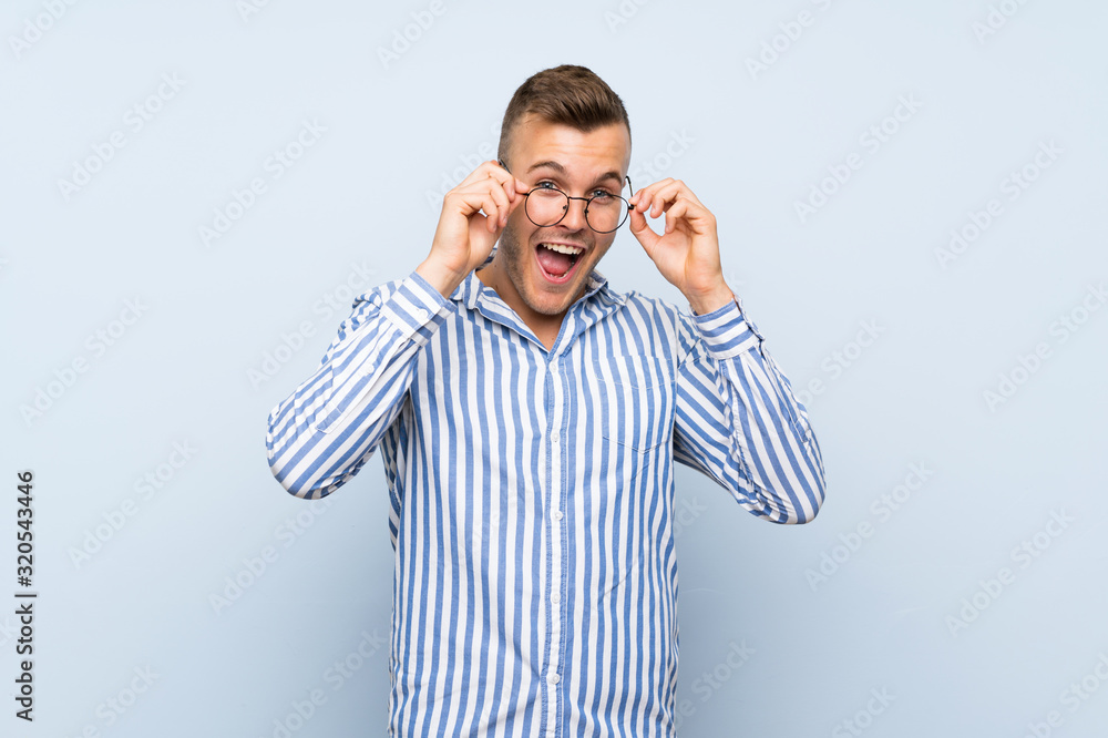 Fototapeta premium Young handsome blonde man over isolated blue background with glasses and surprised