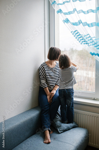 Brunette mother and daughter child spending time at home looking out the window and talking, same clothes and haircuts, full body back view