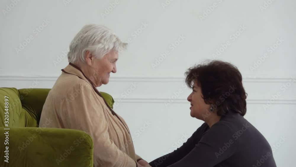 Beautiful middle-aged woman is talking to her elderly mother sitting on a sofa in a white room. Women are smiling Beautiful middle-aged woman is talking to her elderly mother sitting on a sofa in a white room. Women are smiling