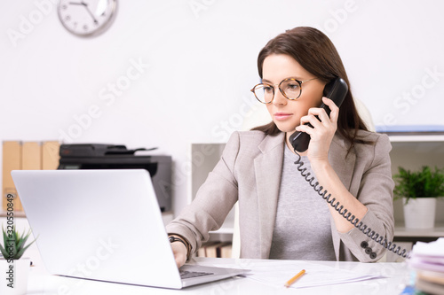 Serious attractive young secretary in glasses answering phone call while searching for information on laptop in office