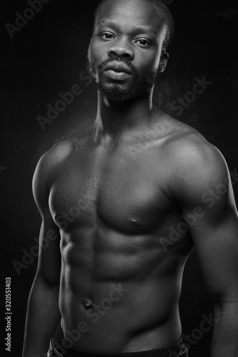 black and white portrait of a handsome black man with naked sports torso looking in the camera on dark background © monchak