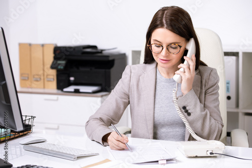 Confident young businesswoman sitting at table and making notes in document while discussing contract content by phone photo