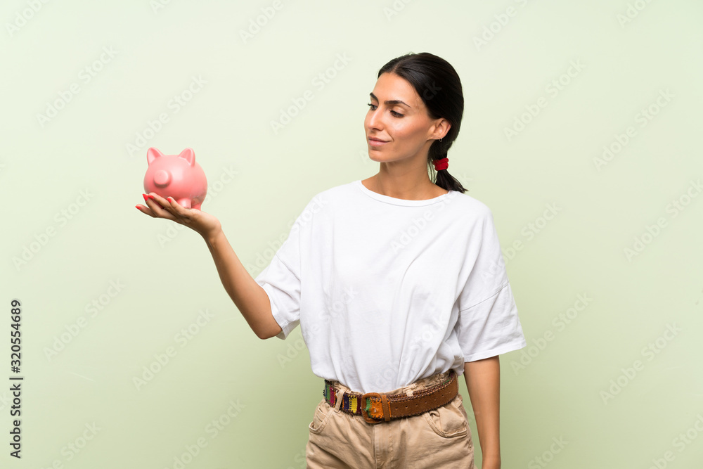 Young woman over isolated green background holding a big piggybank Young woman over isolated green background holding a big piggybank