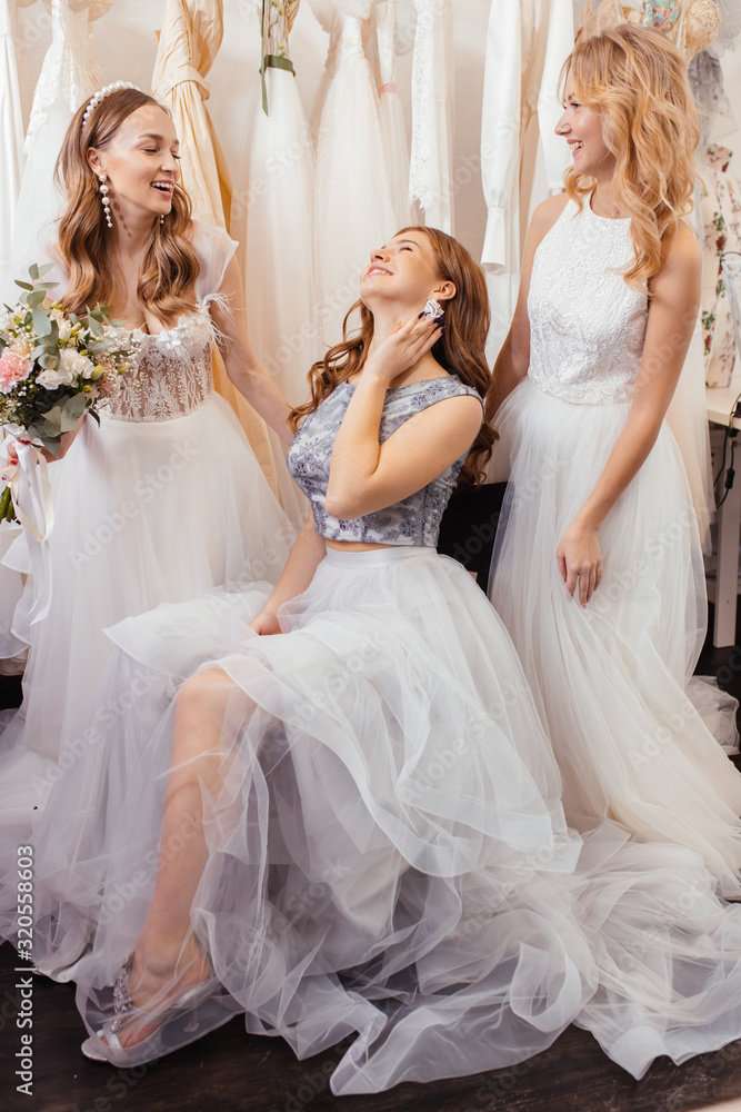 portrait of good-looking three women together in wedding dresses, posing, show new dress made by professional designer tailor, isolated in wedding salon among dresses portrait of good-looking three women together in wedding dresses, posing, show new dress made by professional designer tailor, isolated in wedding salon among dresses