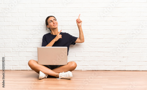 Young woman with a laptop sitting on the floor pointing with the index finger a great idea © luismolinero