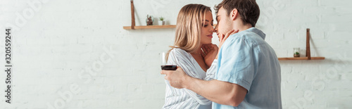 panoramic shot of handsome man holding glass of wine and kissing woman at home