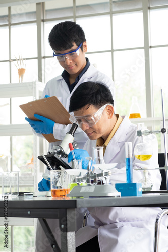 Science, Chemistry, Technology, Biology and Laboratory concept - Asian Senior scientist looks into something on a petri dish while Asian junior scientist taking notes.