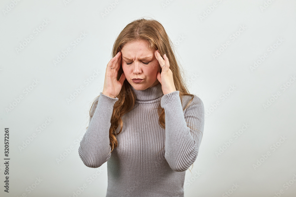 A young beautiful red haired woman in gray sweater with headache holds her hands on head while enduring pain and problems of the disease A young beautiful red haired woman in gray sweater with headache holds her hands on head while enduring pain and problems of the disease