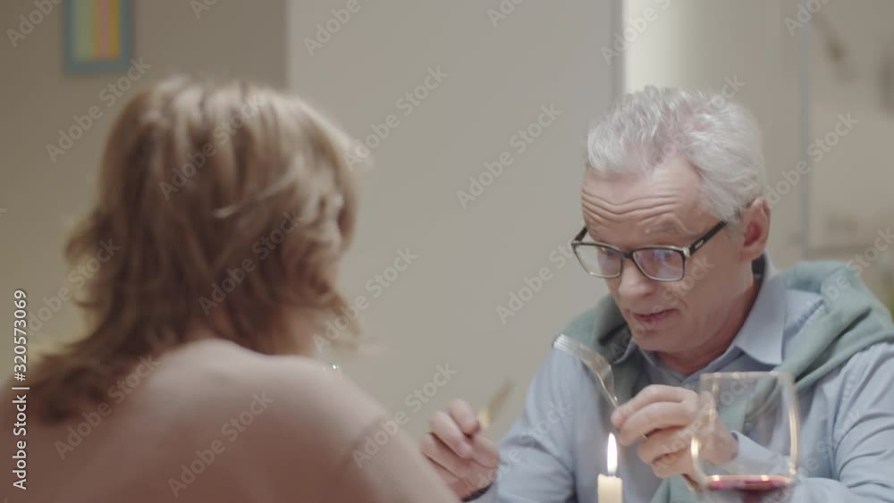 Senior gray-haired man in eyeglasses speaking with wife while sitting together at dining table and eating dinner at home Senior gray-haired man in eyeglasses speaking with wife while sitting together at dining table and eating dinner at home