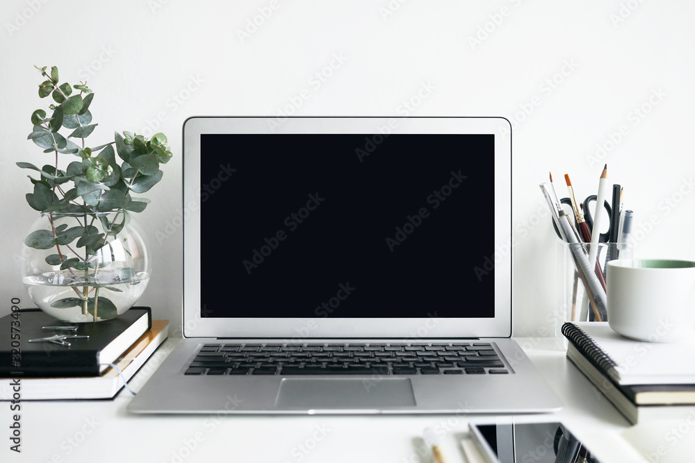Fototapeta premium Horizontal image of modern stylish workplace with open generic portable computer, mobile phone, books, office accessories and decorative plant on white desk. Perfect space for creative work
