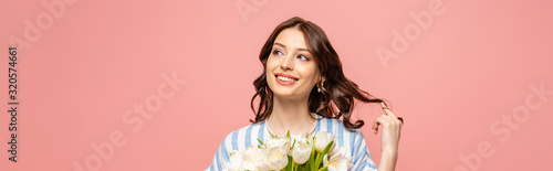 panoramic shot of cheerful girl touching hair while holding bouquet of white tulips isolated on pink