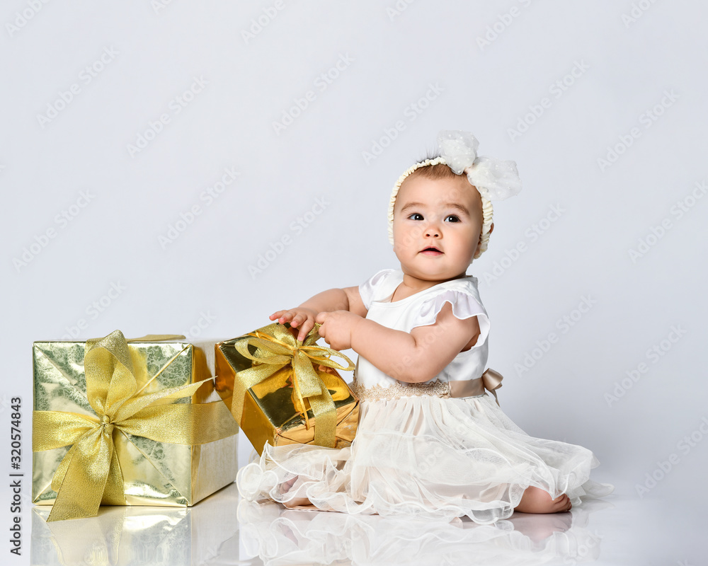Fototapeta premium Baby girl in dress and headband, barefoot. She holding golden gift box, sitting on floor isolated on white. Close up, copy space