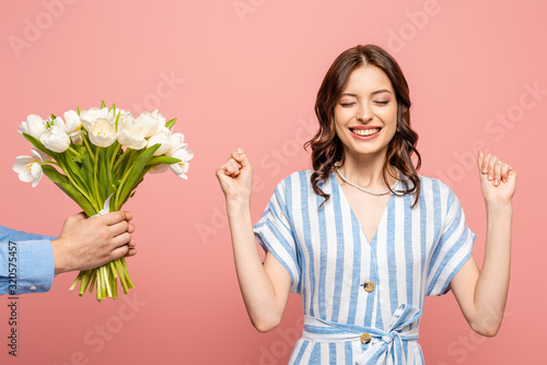 cropped view of man presenting bouquet of white tulips to excited young woman showing winner gesture isolated on pink