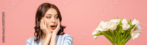 panoramic shot of surprised girl touching face while looking at bouquet of white tulips isolated on pink