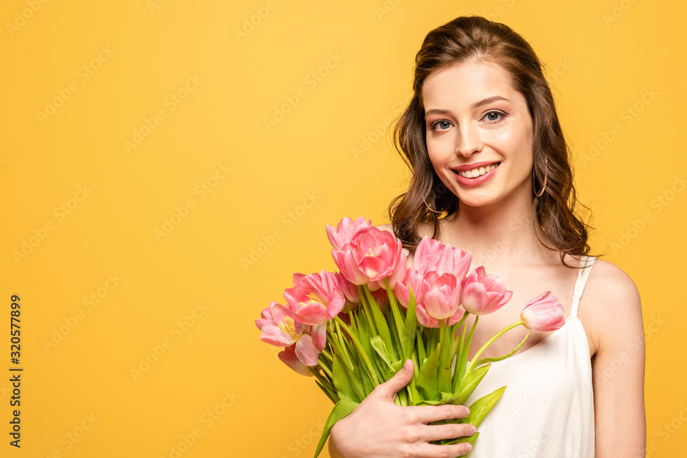 cheerful young woman smiling at camera while holding bouquet of pink tulips isolated on yellow