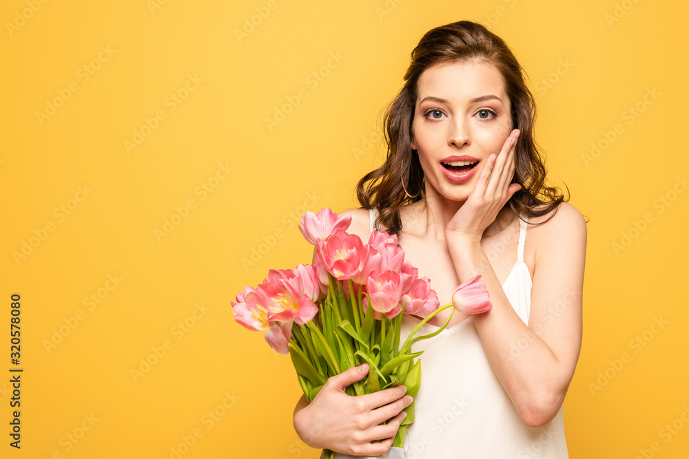 shocked young woman holding bouquet of pink tulips and touching face while looking at camera isolated on yellow