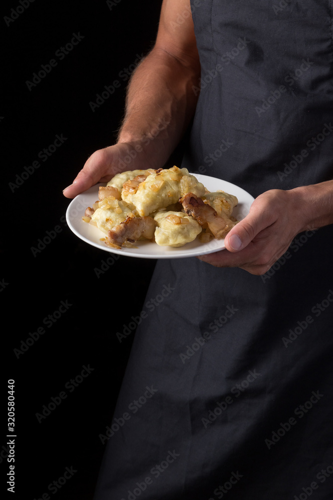 Man holds a plate with varenyky or dumplings, with meat, onions and bacon on a white plate. Pierogi, dumplings served with caramelized salted onion in bowl. Male in a black apron without face. close-u