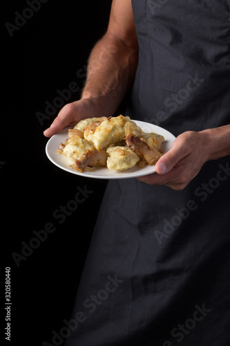 Man holds a plate with varenyky or dumplings, with meat, onions and bacon on a white plate. Pierogi, dumplings served with caramelized salted onion in bowl. Male in a black apron without face. close-u